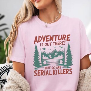 A woman wearing a pink t-shirt with a graphic design that says "Adventure is out there! But so are serial killers".