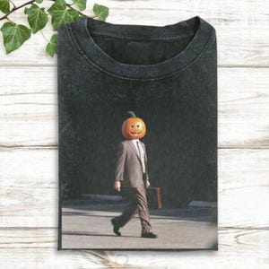 May include: Black t-shirt featuring a surreal image of a person with a carved pumpkin head, wearing a suit and tie, and carrying a briefcase. The person is walking on a crosswalk.