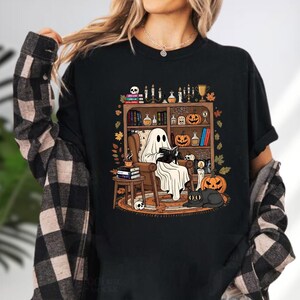 A person wearing a black t-shirt with a graphic design depicting a ghost reading a book in a room filled with Halloween-themed decorations and objects.