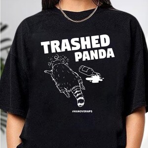 A black t-shirt with a graphic design featuring a raccoon and a spilled liquid, along with the text "TRASHED PANDA" printed on it.