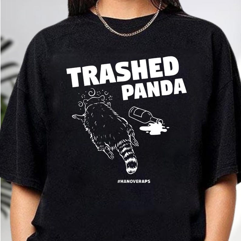 A black t-shirt with a graphic design featuring a raccoon and a spilled liquid, along with the text "TRASHED PANDA" printed on it.