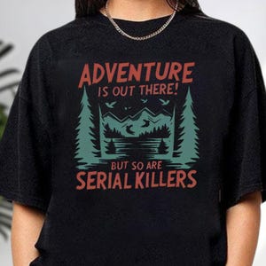 A black t-shirt with a graphic design featuring a forest scene and the text "Adventure is out there! But so are serial killers" printed on it.