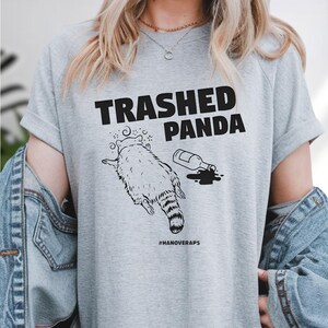 A person wearing a gray t-shirt with a graphic design depicting a raccoon and a bottle of alcohol, along with the text "Trashed Panda #hANoveraps".