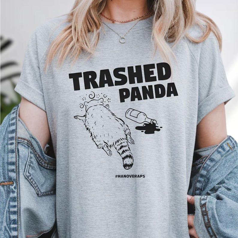 A person wearing a gray t-shirt with a graphic design depicting a raccoon and a bottle of alcohol, along with the text "Trashed Panda #hANoveraps".