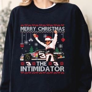 May include: Navy blue sweatshirt with a Christmas design. The graphic features a race car, a driver in a Santa hat, and the text "MERRY CHRISTMAS" and "THE INTIMIDATOR". Festive elements like snowflakes and hearts are also included.