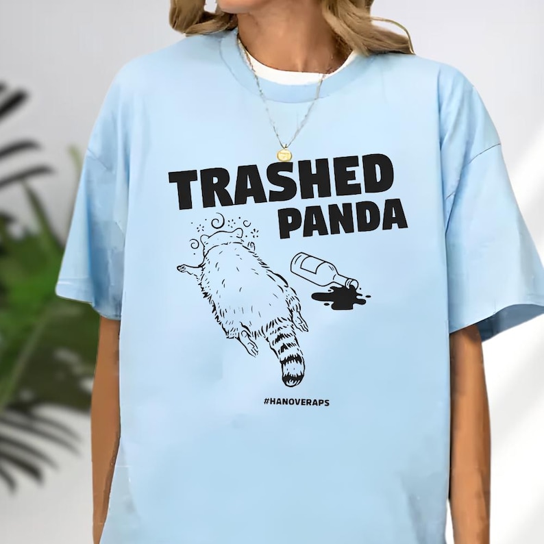 A light blue t-shirt with a graphic design depicting a raccoon and a bottle of alcohol, along with the text "TRASHED PANDA" and "#HANOVERAPS" printed on it.