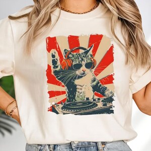 A white t-shirt with a graphic design depicting a cat wearing sunglasses and headphones, playing a turntable.