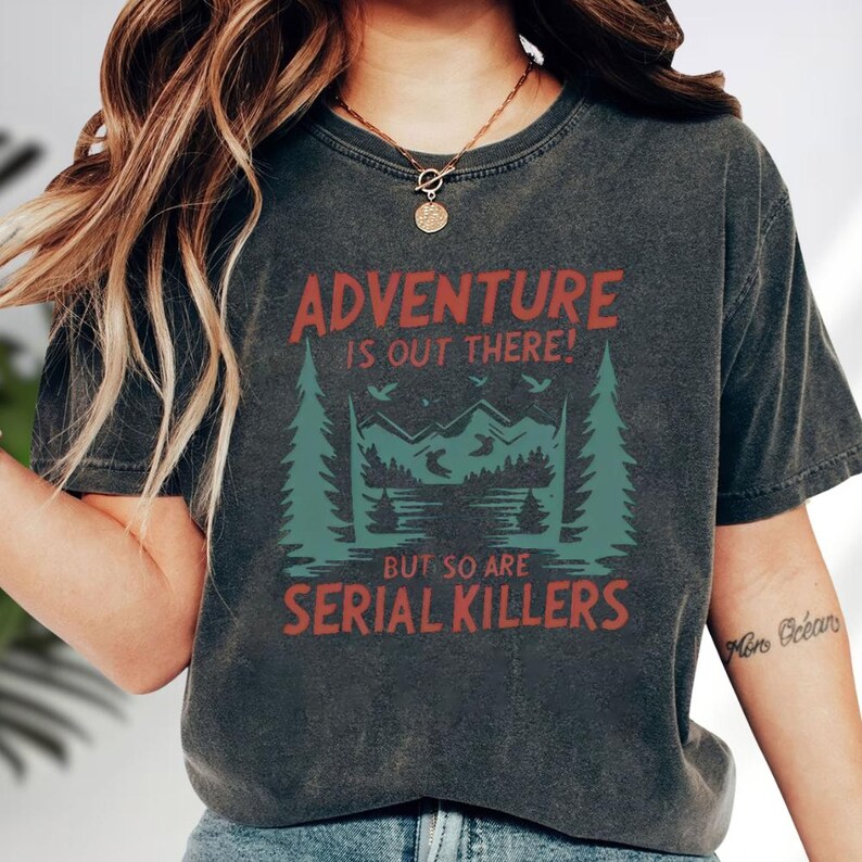 A person wearing a black t-shirt with a graphic design that says "Adventure is out there! But so are serial killers" printed on it.
