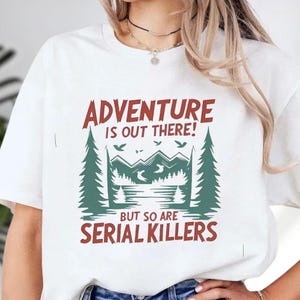 A white t-shirt with a graphic design that says "Adventure is out there! BUT SO ARE SERIAL KILLERS" printed on it.