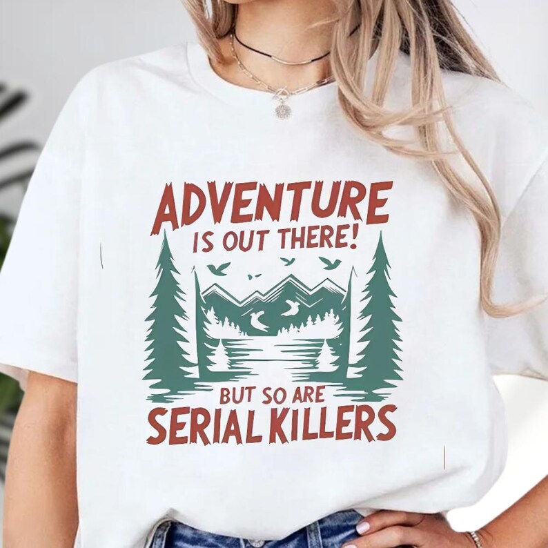 A white t-shirt with a graphic design that says "Adventure is out there! BUT SO ARE SERIAL KILLERS" printed on it.