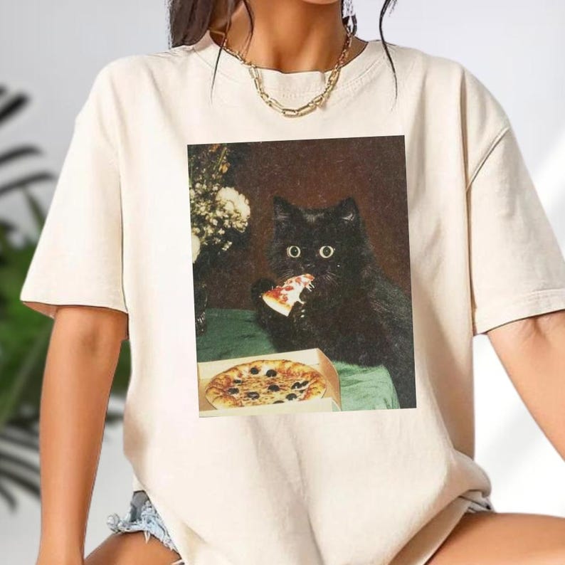 A person wearing a white t-shirt with a black cat sitting on a bed and eating a slice of pizza.