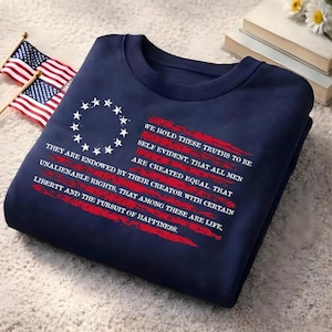 May include: A navy blue sweatshirt featuring a white circular arrangement of thirteen stars and red brushstroke stripes. The text reads: WE HOLD THESE TRUTHS TO BE SELF EVIDENT, THAT ALL MEN ARE CREATED EQUAL. THAT THEY ARE ENDOWED BY THEIR CREATOR WITH CERTAIN UNALIENABLE RIGHTS, THAT AMONG THESE ARE LIFE, LIBERTY AND THE PURSUIT OF HAPPINESS.