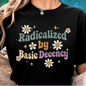 May include: Black t-shirt with the text "Radicalized by Basic Decency" in a retro font. The design includes daisy and star graphics in various colors. A casual, everyday wear tee.