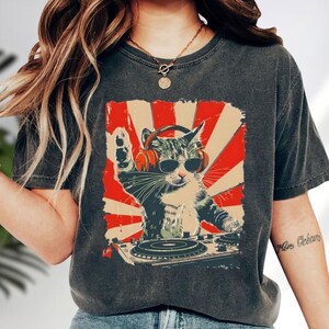A person wearing a gray t-shirt with a graphic design of a cat wearing headphones and sunglasses, playing a turntable, and surrounded by a red and white sunburst background.