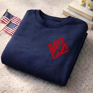 May include: Navy blue sweatshirt with a red logo on the chest. The logo is a stylized design. Two small American flags are in the background.