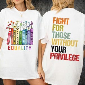 May include: White t-shirt with a colorful graphic on the front featuring the words "LOVE, PEACE, INCLUSION, KINDNESS, CHANGE, DIVERSITY, UNITY, COMPASSION, JUSTICE" and the word "EQUALITY". The back of the shirt says "FIGHT FOR THOSE WITHOUT YOUR PRIVILEGE".