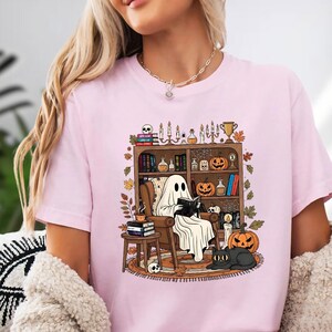 A woman wearing a pink t-shirt with a graphic design depicting a ghost reading a book in a room filled with Halloween decorations and objects.