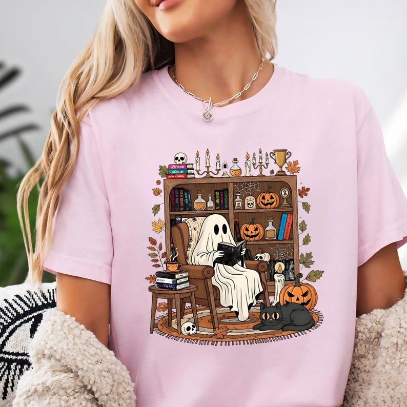 A woman wearing a pink t-shirt with a graphic design depicting a ghost reading a book in a room filled with Halloween decorations and objects.
