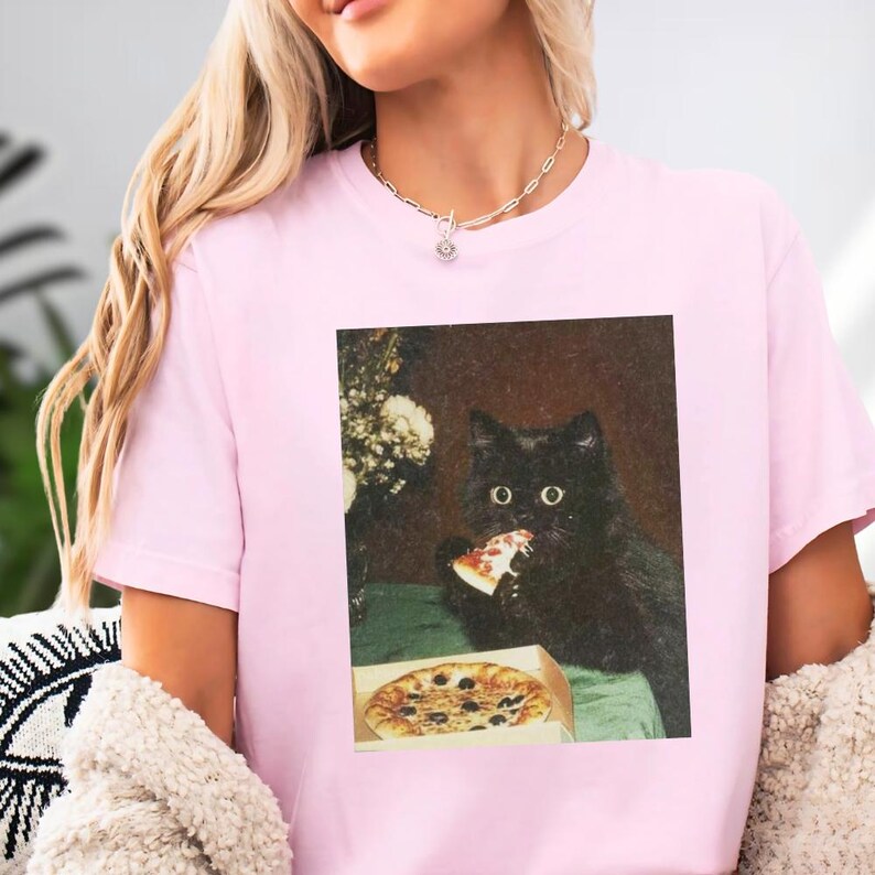 A woman wearing a pink t-shirt with a black cat eating pizza.
