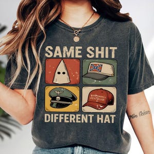 May include: Dark gray t-shirt with the text "SAME SHIT DIFFERENT HAT" and four images of different hats. The hats include a white hood, a Confederate flag hat, a military hat, and a "Make America Great Again" hat.
