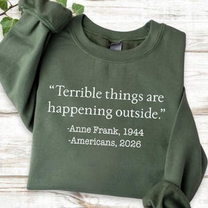 May include: An olive green sweatshirt with the quote "Terrible things are happening outside." in white text. The text also includes "-Anne Frank, 1944" and "-Americans, 2026."