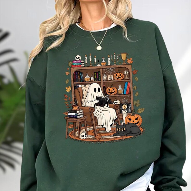 A green sweatshirt with a Halloween-themed graphic depicting a ghost sitting in a chair surrounded by various Halloween-related items, such as pumpkins, bats, and candles.