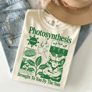 May include: A cream-colored t-shirt featuring a green graphic print of a cat and a photosynthesis diagram. The text reads Photosynthesis, Sunlight, water, carbon dioxide, sugar, O2, and Brought To You By The Sun. The shirt has a Comfort Colors label.