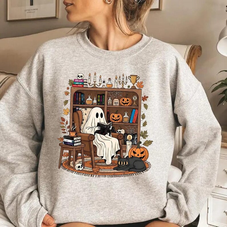 A woman wearing a gray sweatshirt with a Halloween-themed graphic design depicting a ghost sitting in a chair surrounded by various Halloween-related items.