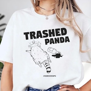 A white t-shirt with a graphic design depicting a panda with a bottle of liquid, accompanied by the text "TRASHED PANDA" and "#HAINOVERAPS".