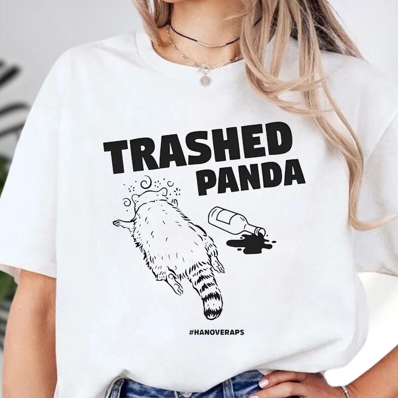 A white t-shirt with a graphic design depicting a panda with a bottle of liquid, accompanied by the text "TRASHED PANDA" and "#HAINOVERAPS".