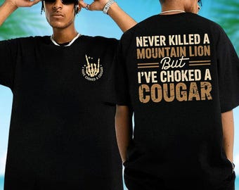 May include: A black short-sleeved t-shirt featuring a small graphic on the front chest of a skeleton hand with the text "I HAVE CHOKED A COUGAR". The back displays the text "NEVER KILLED A MOUNTAIN LION But I'VE CHOKED A COUGAR" in tan lettering.