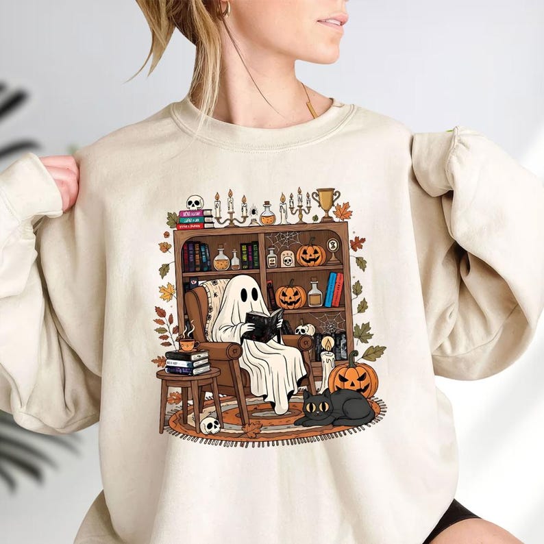 A woman wearing a white sweatshirt with a graphic design depicting a ghost sitting in a chair surrounded by Halloween-themed items such as pumpkins, books, and candles.