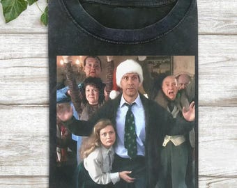 May include: Black t-shirt featuring a movie scene with a man wearing a Santa hat and a green tie, surrounded by several people. The image is a still from a Christmas movie. The shirt has a crew neck.