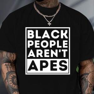 May include: Black t-shirt with a white rectangular graphic. The graphic contains the text "BLACK PEOPLE AREN'T APES" in a bold, sans-serif font. The shirt is a solid black color.