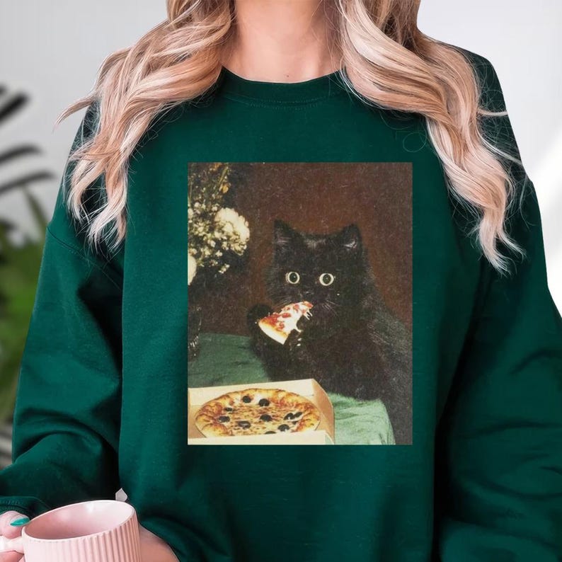A person wearing a green sweater with a printed image of a black cat eating pizza.