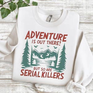 A white sweatshirt with a graphic design on it, featuring a forest scene with mountains and pine trees, along with the text "Adventure is out there! BUT SO ARE SERIAL KILLERS".