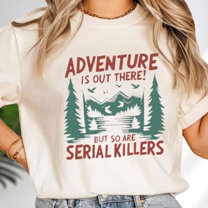 A woman wearing a white t-shirt with a graphic design that says "Adventure is out there! But so are serial killers" printed on it.