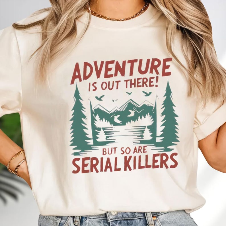 A woman wearing a white t-shirt with a graphic design that says "Adventure is out there! But so are serial killers" printed on it.