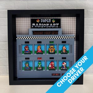 May include: A framed pixel art piece of the Super Mario Kart character selection screen. The screen shows the different characters you can choose from, including Mario, Luigi, Peach, Toad, Donkey Kong, and Bowser. The frame is black and the background is gray.