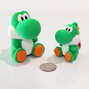 May include: Two green and white Yoshi plush toys, one large and one small, sit side-by-side. The toys have orange feet and a knitted texture. A US quarter is placed between them for size comparison.