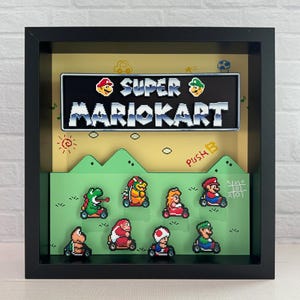 May include: A black framed shadow box with a 3D diorama of the Super Mario Kart video game. The diorama features characters from the game, including Mario, Luigi, Toad, and Yoshi, driving their karts on a green track. The box also includes a title card with the game's logo.