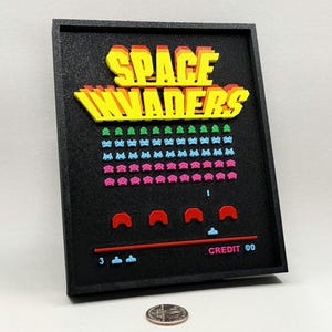 May include: A black framed wall art piece featuring the Space Invaders arcade game. The artwork displays the game's title in yellow and red, along with colorful pixelated alien invaders and game elements. The credit is 00.