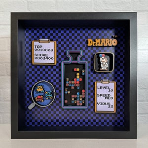 May include: A framed 3D diorama of the classic Nintendo game Dr. Mario. The diorama features a blue and black checkered background, a clear pill bottle with colorful pills, a scorecard, a level card, and a character from the game.