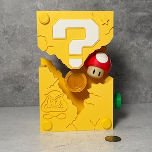 May include: A yellow question mark block from the Super Mario video game series. The block features a white question mark, a red and white mushroom, a gold coin, and a Goomba character. The block has a cracked design and a green button.