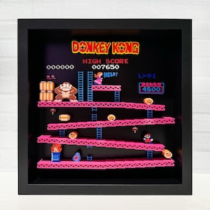 May include: A framed pixel art recreation of the classic Donkey Kong arcade game. The image shows Donkey Kong, Mario, and Pauline in a pixelated style. The game's score, level, and bonus are displayed on the screen. The frame is black and the background is black.