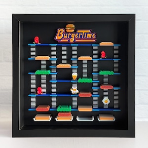 May include: A black framed shadow box with a black background and a white and yellow "BurgerTime" logo. The box contains a variety of colorful pixelated food and character figures, including burgers, fries, eggs, and a chef.