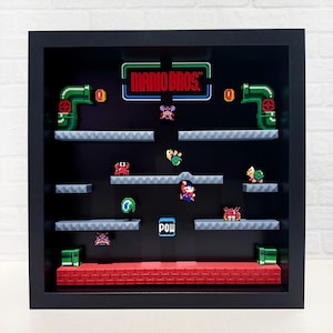 May include: A framed 3D pixel art scene of the classic video game 'Mario Bros.' The scene features Mario, a Goomba, a Koopa Troopa, and other iconic characters from the game. The frame is black and the background is black with gray platforms. The text 'MARIO BROS.' is displayed in a neon green and blue font.