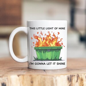 Op de afbeelding: Witte keramische mok met een brandende groene vuilnisbak en de tekst "This little light of mine I'm gonna let it shine".