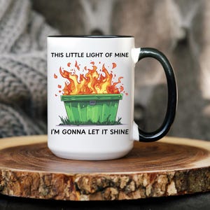 Op de afbeelding: Witte keramische mok met een zwarte rand en handvat. De mok heeft een groene vuilnisbak met vlammen erop en de tekst "This little light of mine I'm gonna let it shine".