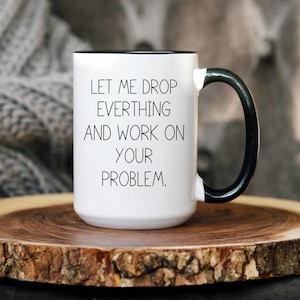 May include: White ceramic coffee mug with a black rim and handle. The mug has black text that reads "Let me drop everything and work on your problem."